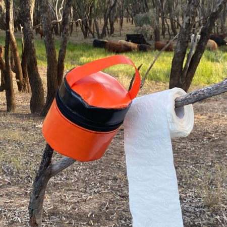 PVC toilet roll holder hanging on a tree branch with a roll of toilet paper, designed for camping and rugged outdoor use.