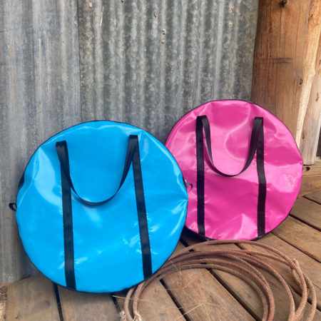 “Pink and sky blue Dust & Iron roping bag with black handles leaning against a tree with lasso rope on red dirt”