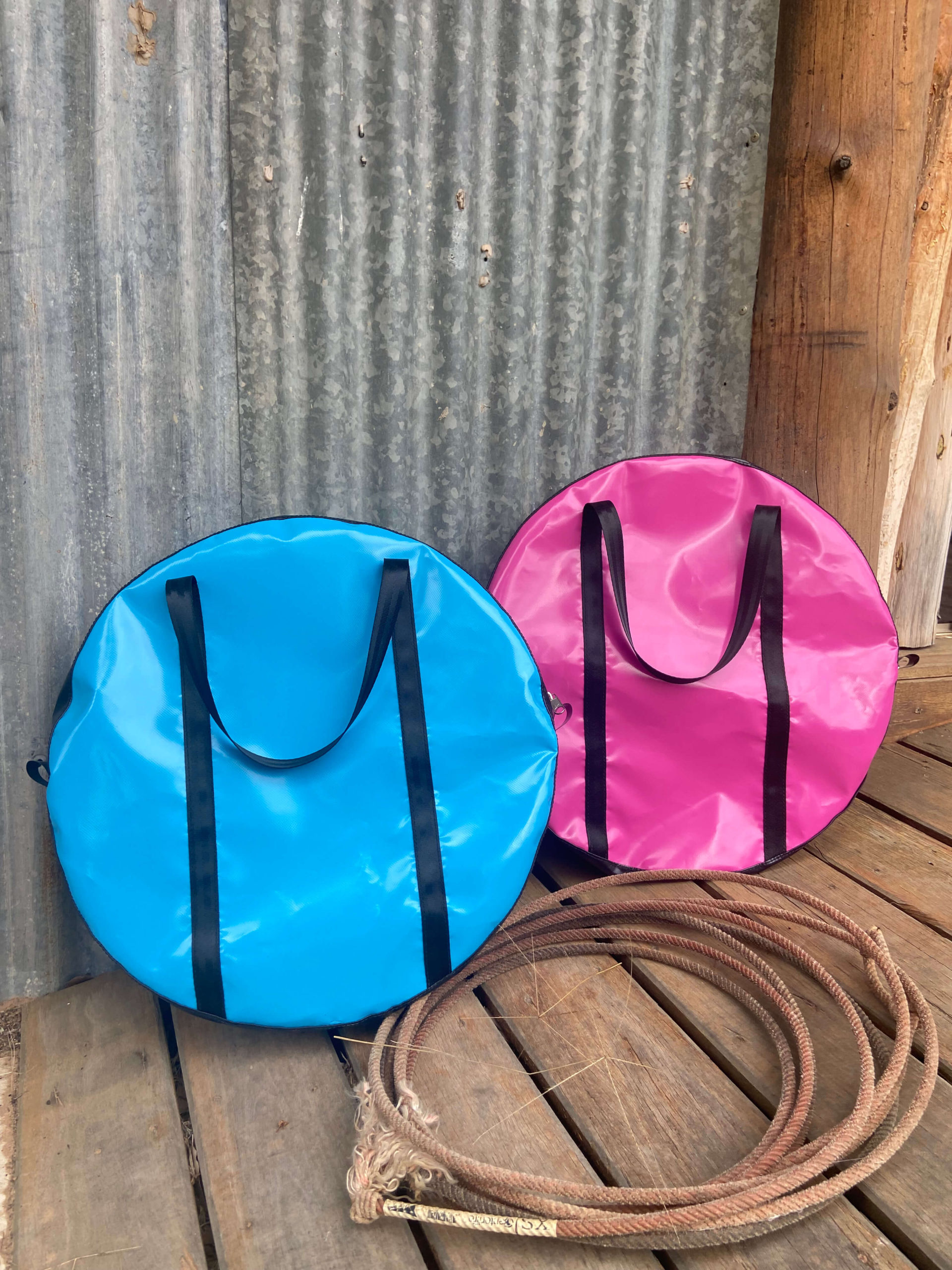 “Pink and sky blue Dust & Iron roping bag with black handles leaning against a tree with lasso rope on red dirt”
