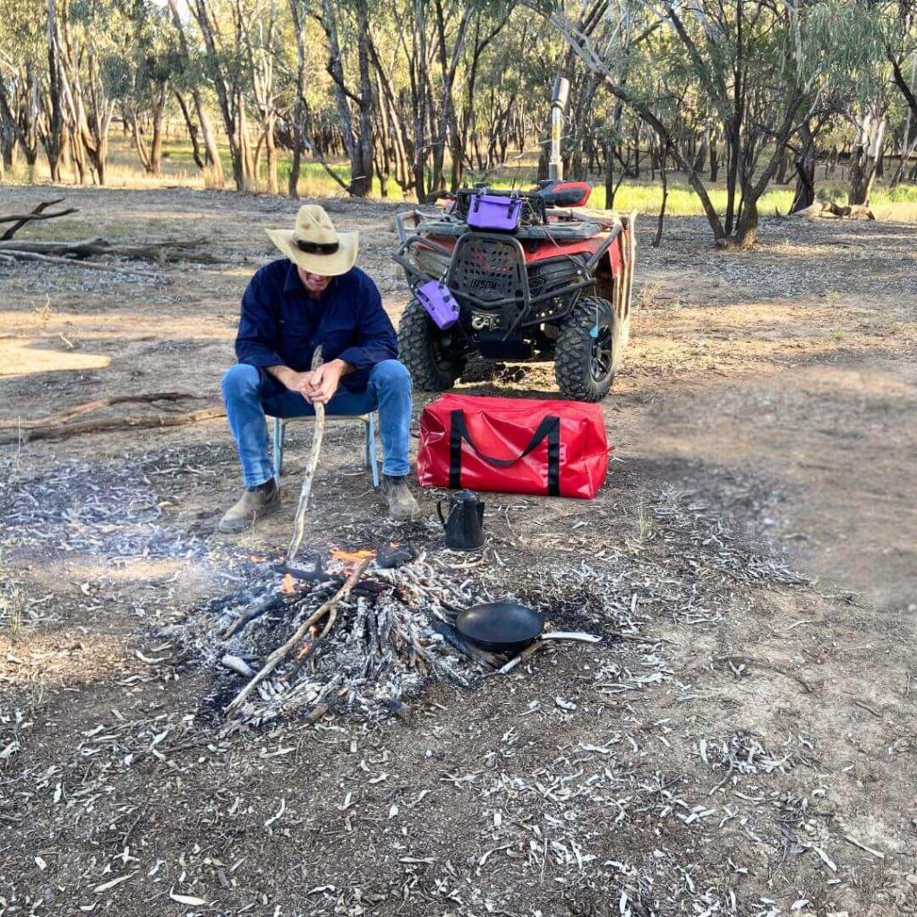 Person sitting by a campfire in the bush with a red Dust & Iron duffel bag and an ATV in the background.