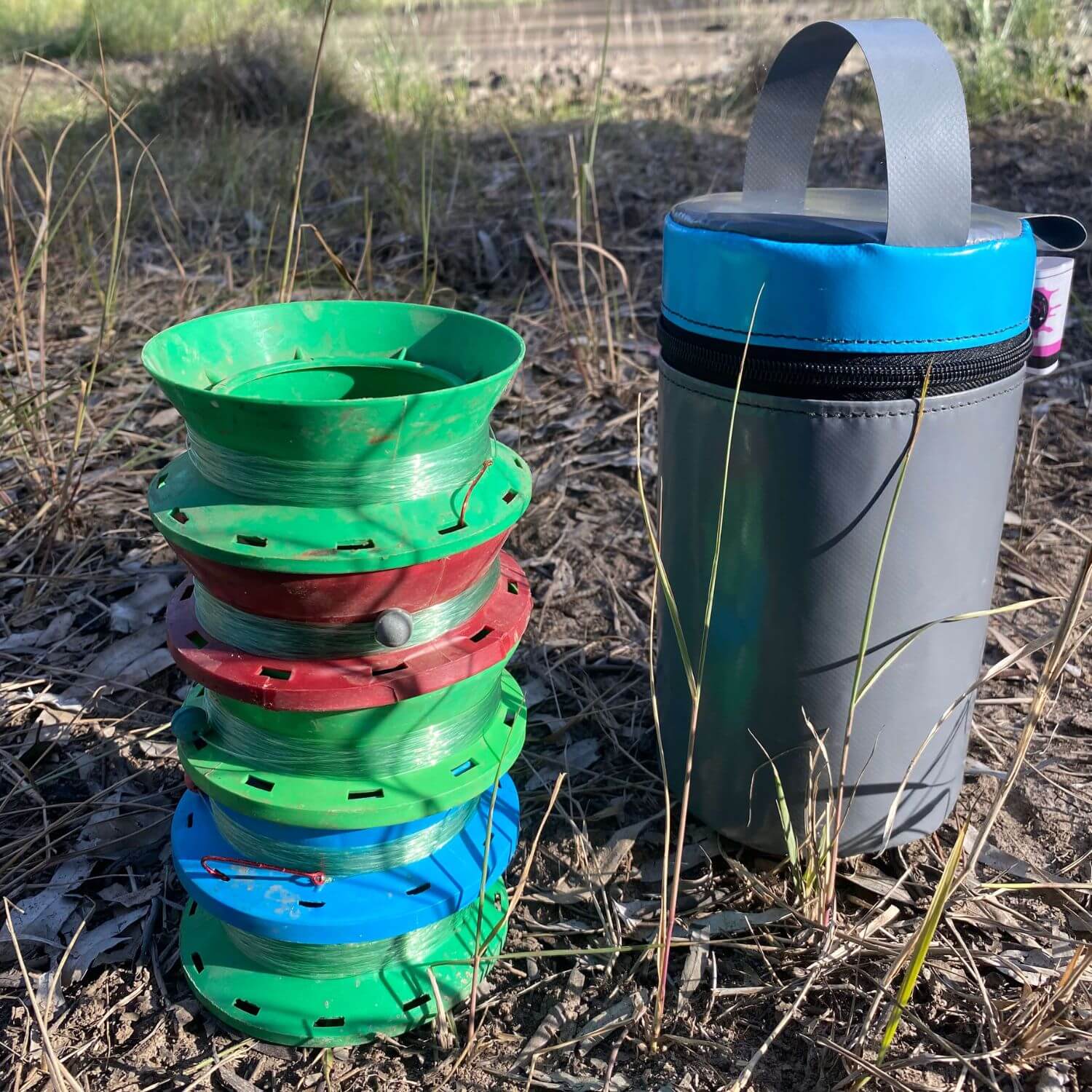 Stacked plastic hand fishing reels in red, green and blue placed next to a grey PVC reel holder bag with a blue lid, photographed outdoors on grass.